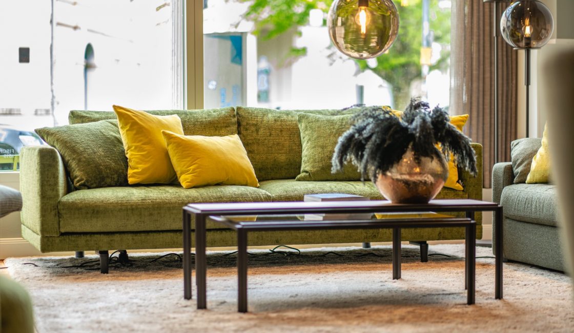 Ein gemütliches Wohnzimmer mit einem grünen Sofa und gelben Kissen. Davor steht ein dunkler Couchtisch mit einer dekorativen Vase darauf. Große Fenster dahinter lassen Tageslicht herein und rahmen die belaubten Zweige eines Baumes draußen ein. Ein beiger Teppich bedeckt den Boden. - Raumausstattung Warnken Oldenburg & Bad Zwischenahn