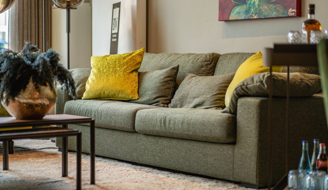 Ein gemütliches Wohnzimmer verfügt über ein grünes Sofa mit gelben Akzentkissen, einen modernen Couchtisch aus Holz mit einer dekorativen Pflanze, einen flauschigen beigen Teppich und Kunstwerke an den Wänden. Eine Stehlampe und Regale mit verschiedenen Objekten runden das Ambiente ab. - Raumausstattung Warnken Oldenburg & Bad Zwischenahn